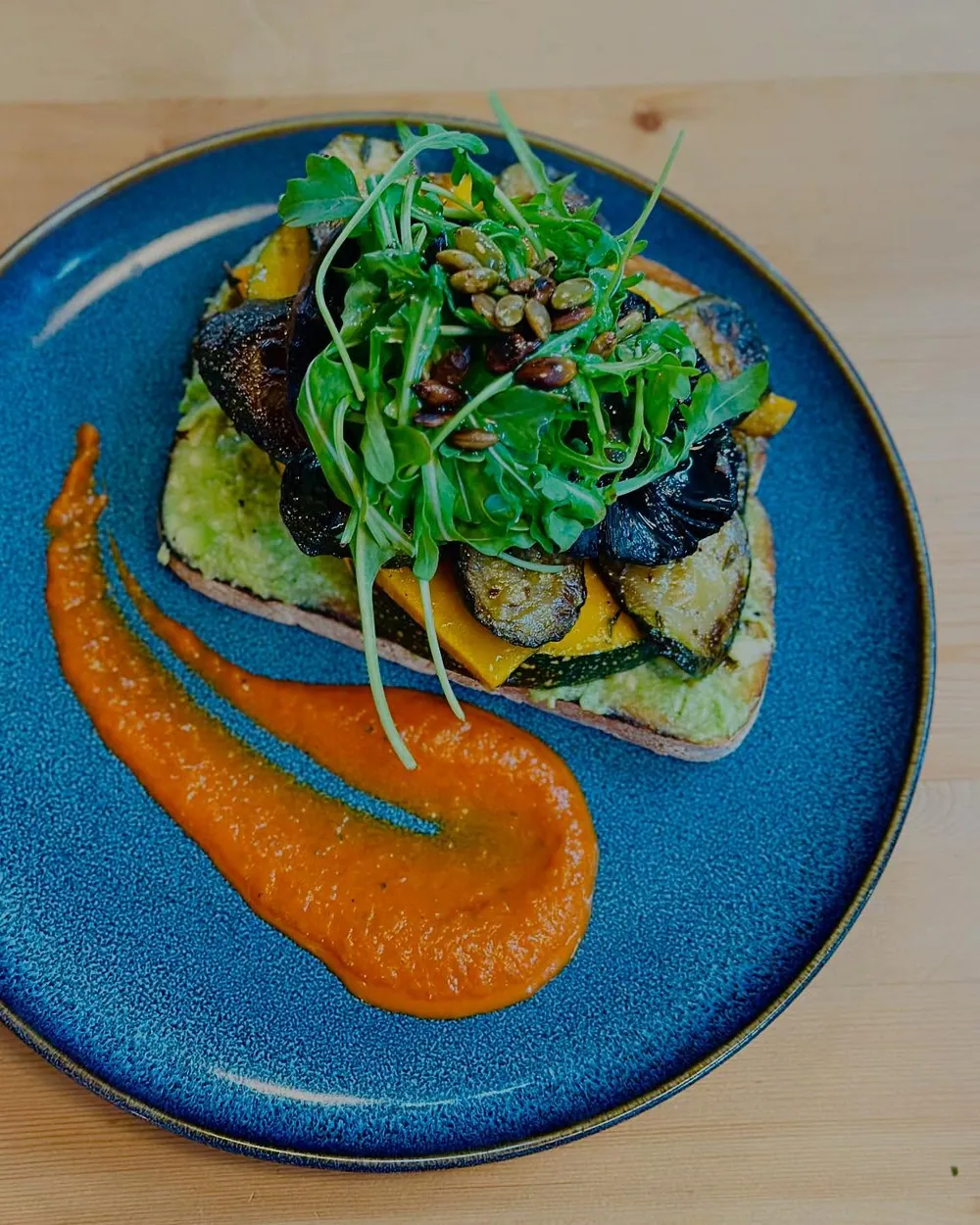 Avocado, greens and roast vegetables on sourdough with a capsicum coulis, on a deep blue plate
